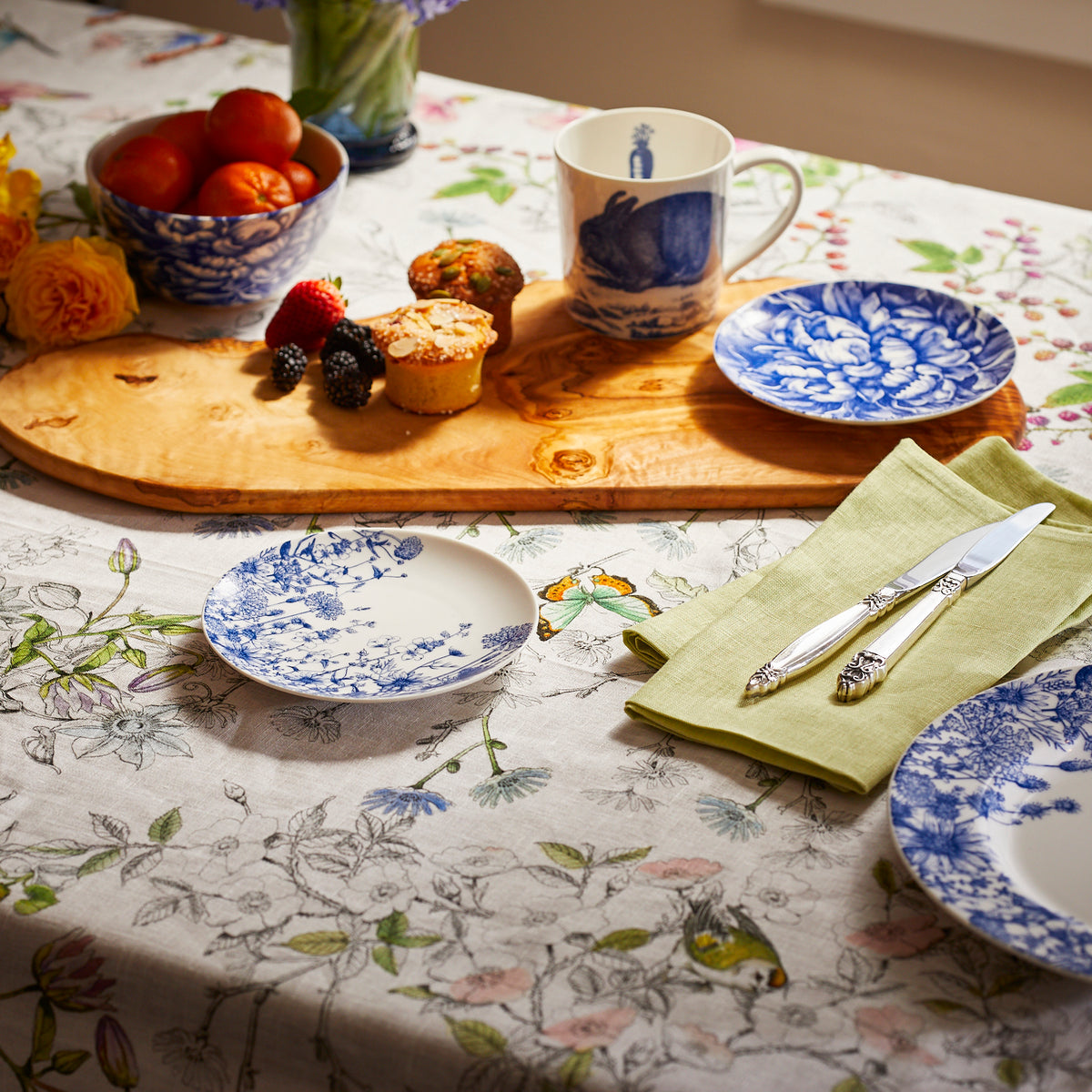 A floral tablecloth with a wooden board displaying muffins, berries, and persimmons. Nearby are a high-fired porcelain Bunnies Mug from Caskata Artisanal Home with a blue design, a matching plate, a yellow napkin, and a set of fork and knife.