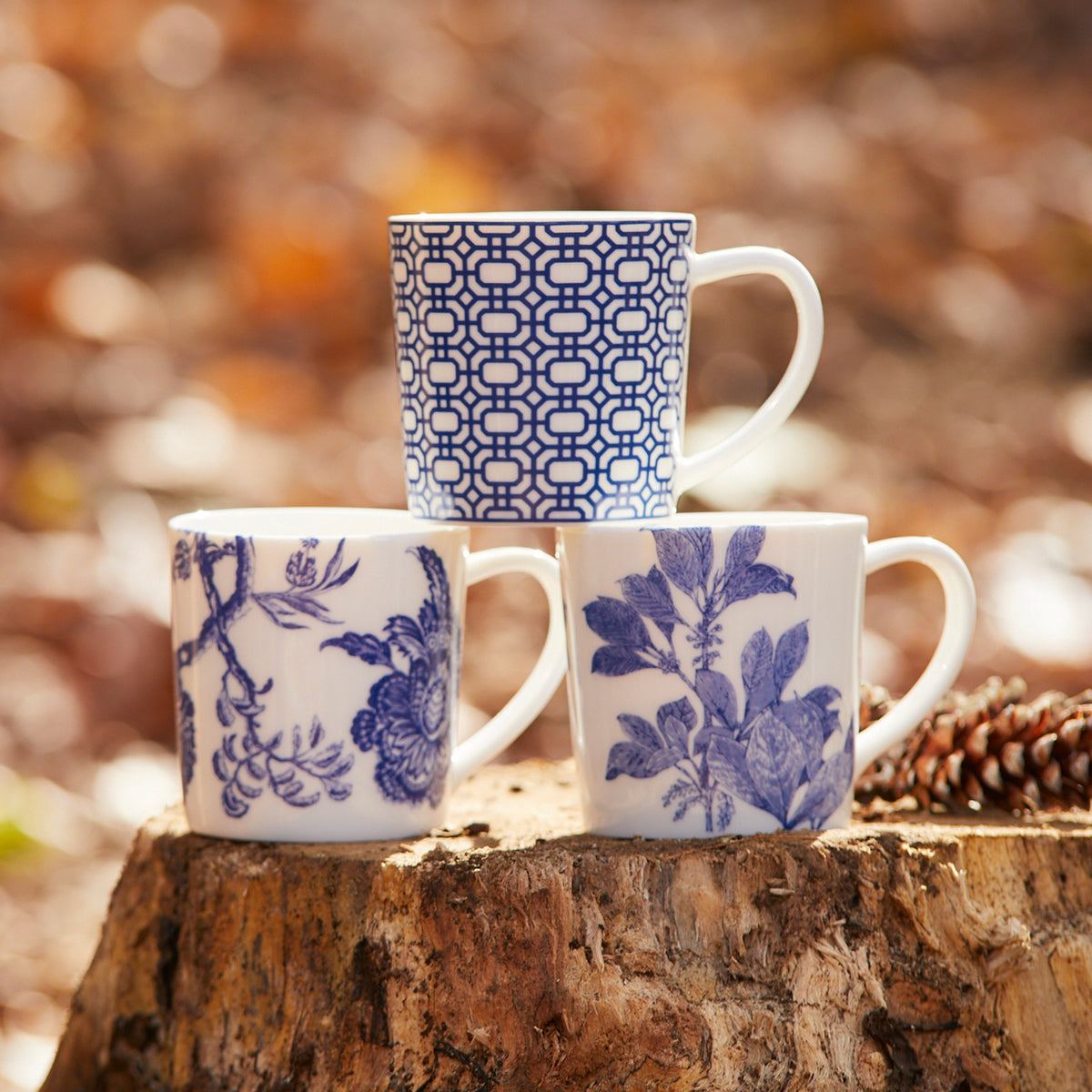 Three creamy white Newport Garden Gate Mugs by Caskata Artisanal Home with blue patterns sit on a tree stump outdoors. One features a geometric design, while the other two boast floral patterns. All are dishwasher and microwave safe.