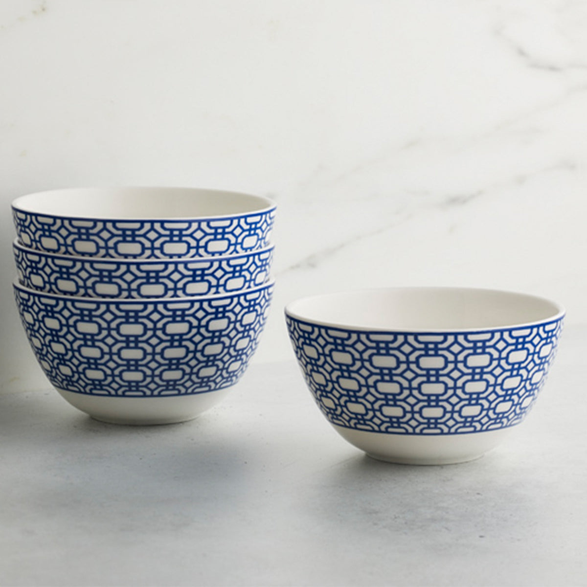 Four premium porcelain bowls with blue geometric patterns sit stacked, while one matching Newport Cereal Bowl from Caskata Artisanal Home is placed beside the stack, on a gray surface near a white marble background.