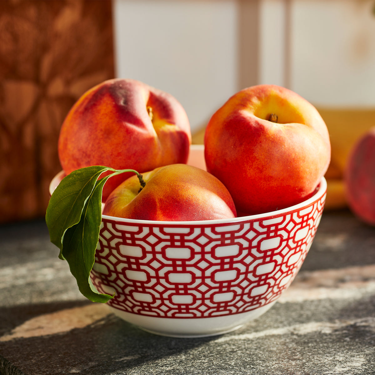 A Caskata Newport Crimson Cereal Bowl with a red geometric pattern holds four peaches, one with a leaf attached. The bowl is placed on a gray stone surface, reminiscent of the timeless elegance found at the Newport Garden Gate.