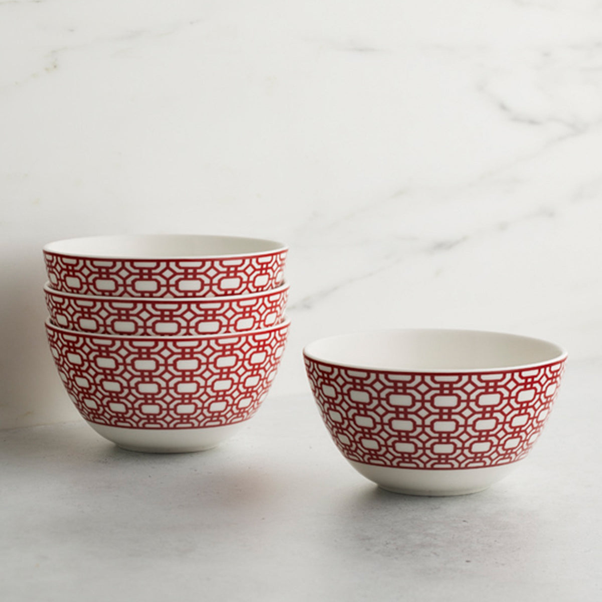 Four white porcelain bowls with a red geometric pattern, three stacked and one placed beside the stack, on a gray surface with a marbled white background. Perfect for your morning cereal, these Newport Crimson Cereal Bowls by Caskata add a touch of elegance to any kitchen setting.