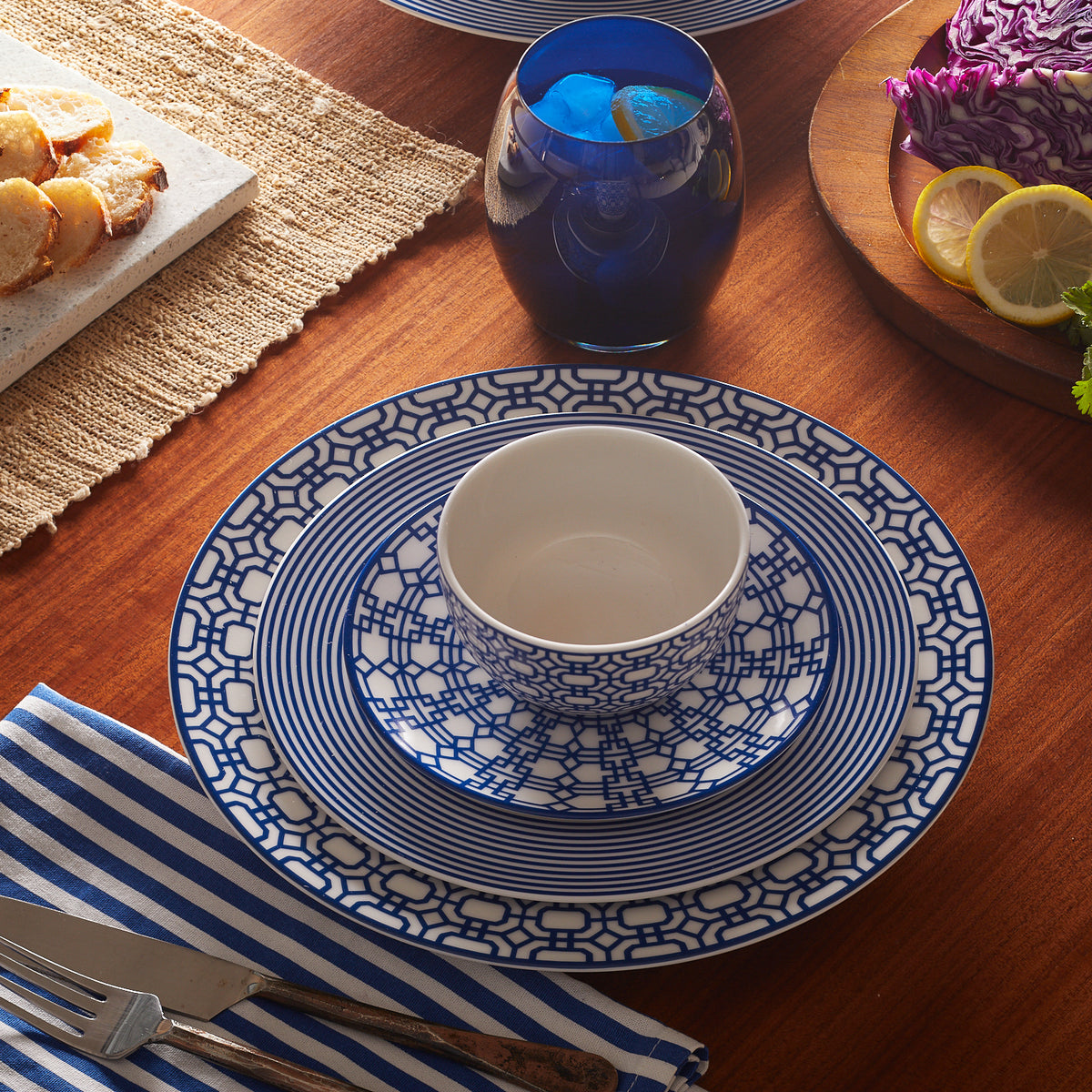 A dining table set with a blue and white patterned bowl, plate, and Newport Small Plates featuring an interlocking lattice pattern, a blue glass with a beverage, bread slices on a cutting board, a striped napkin, knife, and fork. The premium porcelain evokes the charm of Newport Garden Gate from Caskata Artisanal Home.