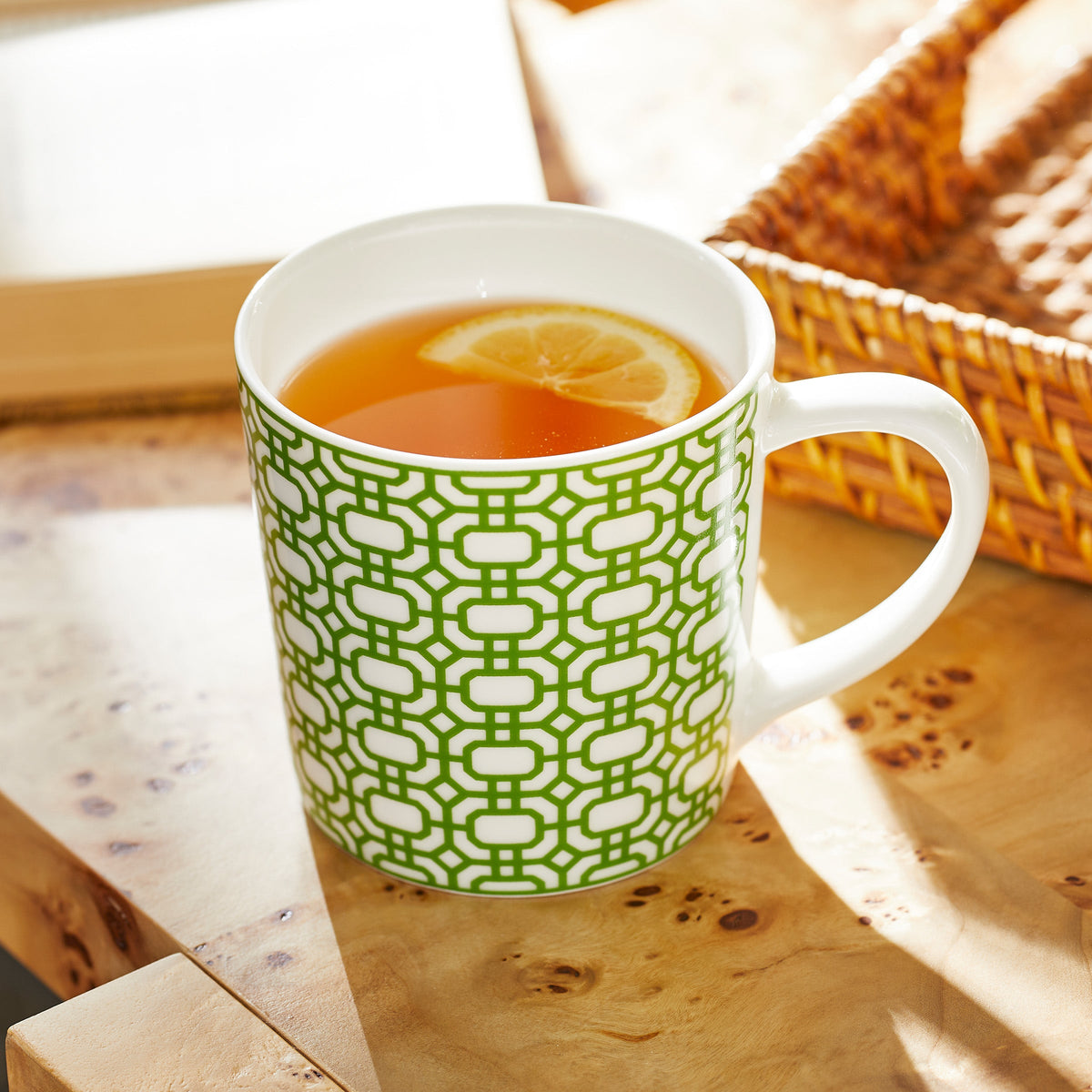 A Newport Garden Gate Verde Mug from Caskata Artisanal Home, filled with light brown tea and a lemon slice, sits on a wooden surface near a wicker basket.