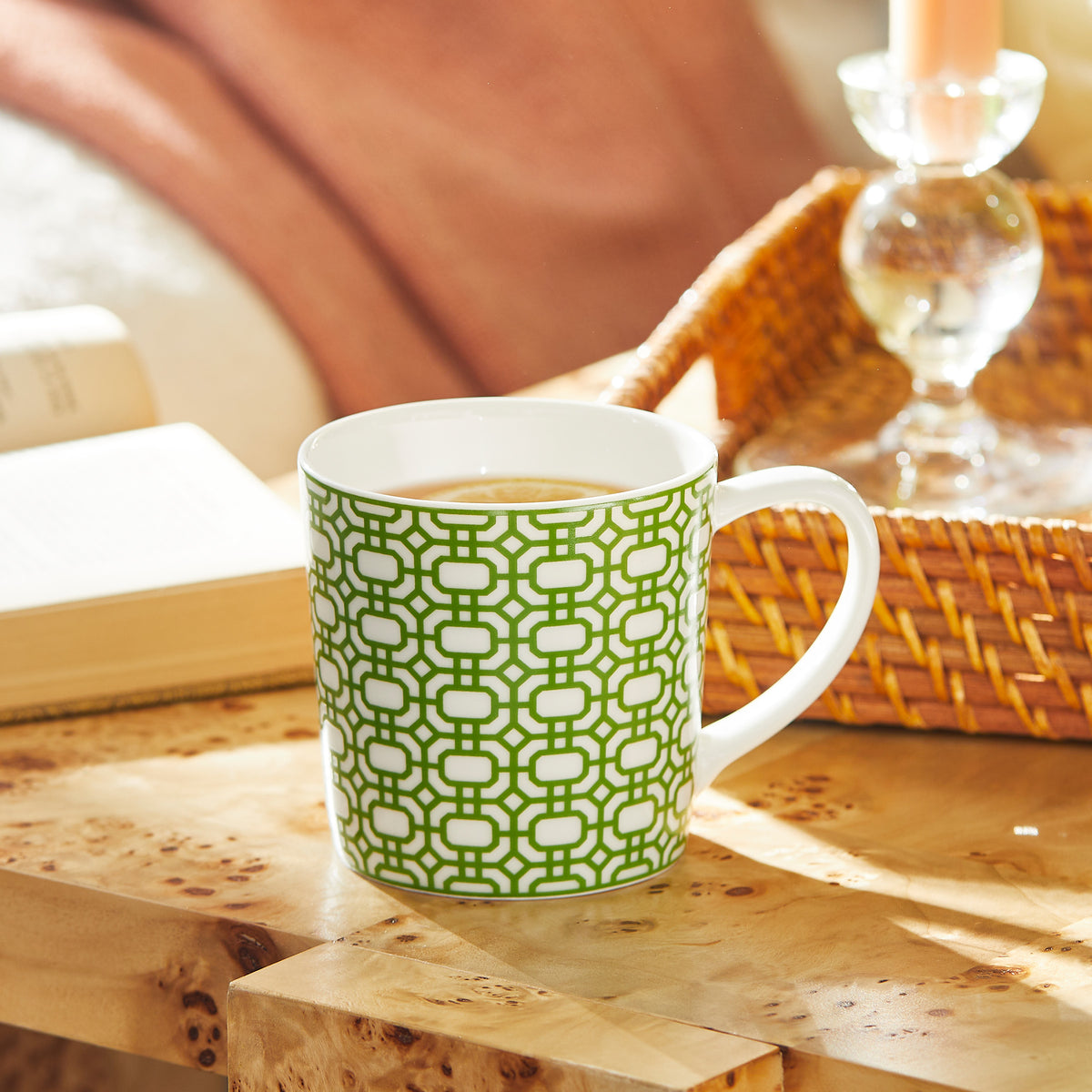 A Caskata Artisanal Home Newport Garden Gate Verde Mug filled with a hot beverage rests on a wooden table beside an open book and a woven basket containing a lit candle.