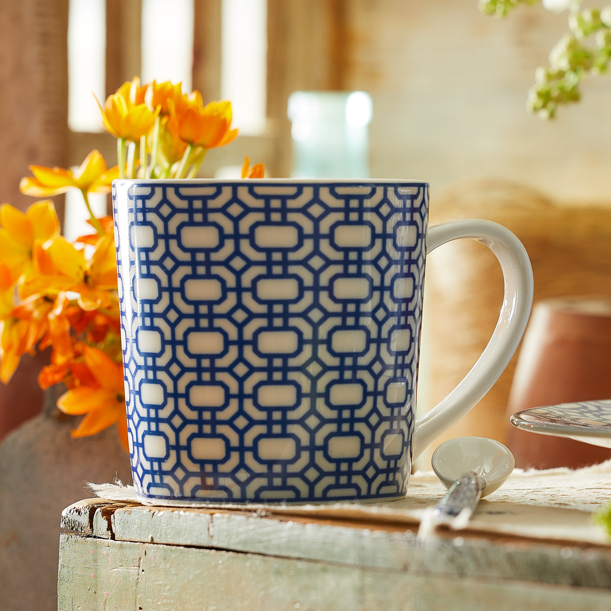 A Newport Garden Gate Mug made by Caskata Artisanal Home, with its blue and white patterned graphic design made of creamy white porcelain, sits on a wooden surface next to a teaspoon and orange flowers, blending charm with practicality as it is both dishwasher and microwave safe.
