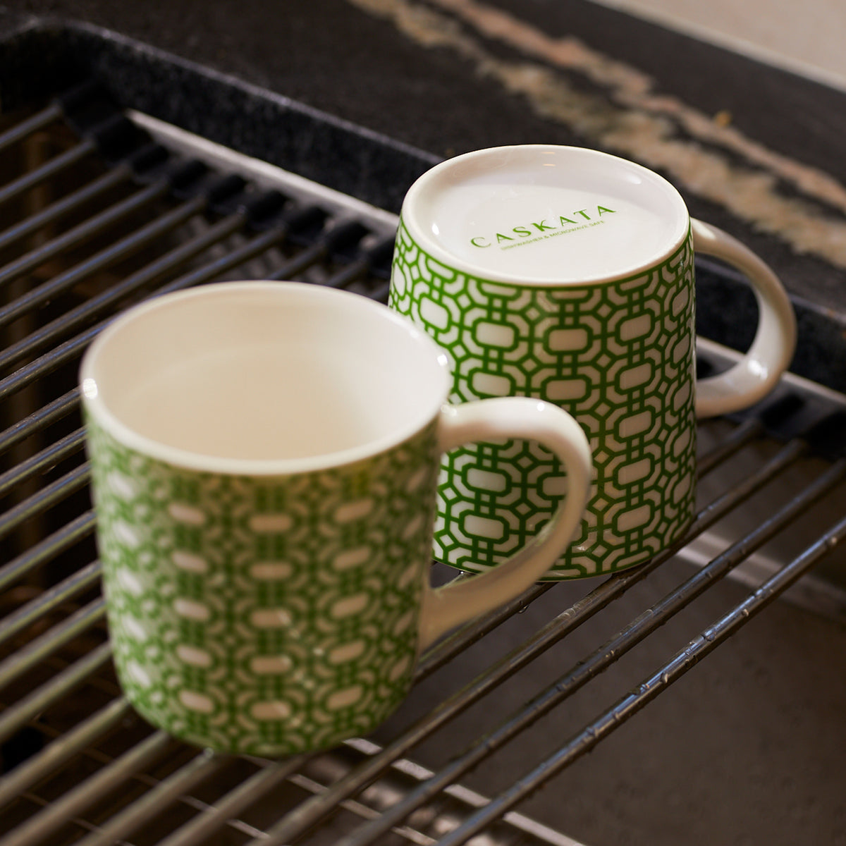 Two green and white chainlink patterned mugs, one right side up and one upside down with &quot;Caskata Artisanal Home&quot; on the base, are placed on a drying rack.