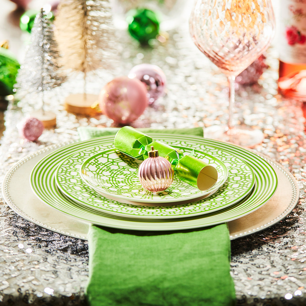 Festive table setting with Newport Stripe Verde Rimmed Dinner Plate by Caskata Artisanal Home, a green napkin, Christmas cracker, and pink ornament on a sparkling tablecloth, surrounded by holiday decorations.