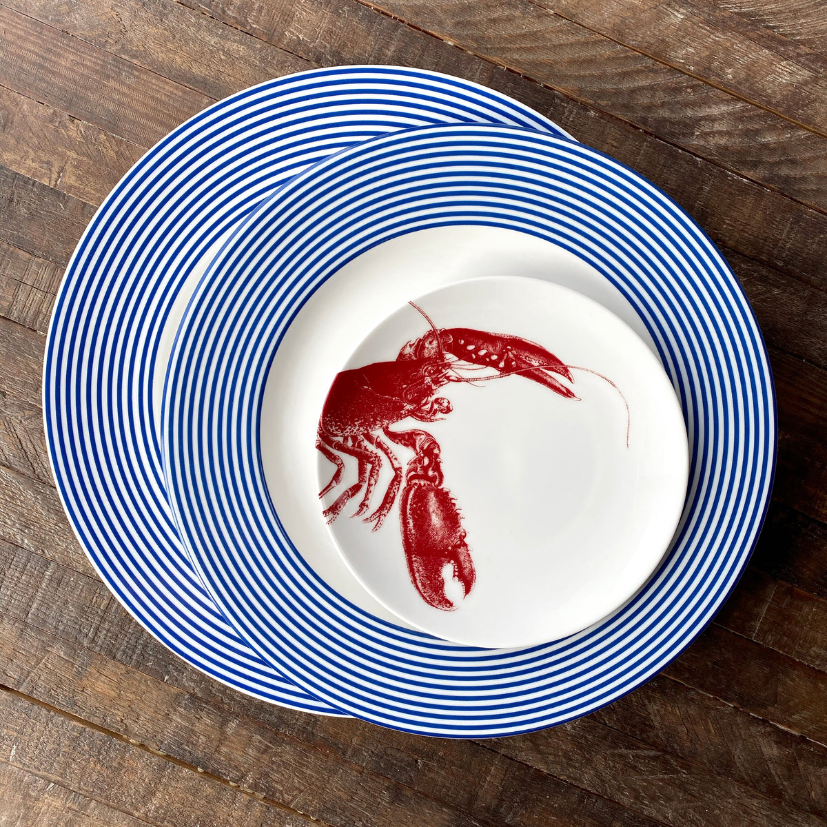 Three high-fired porcelain plates with Newport Stripe patterns are stacked on a wooden surface. The top plate, serving as a Newport Rimmed Charger Plate by Caskata Artisanal Home, features a red lobster illustration in the center.