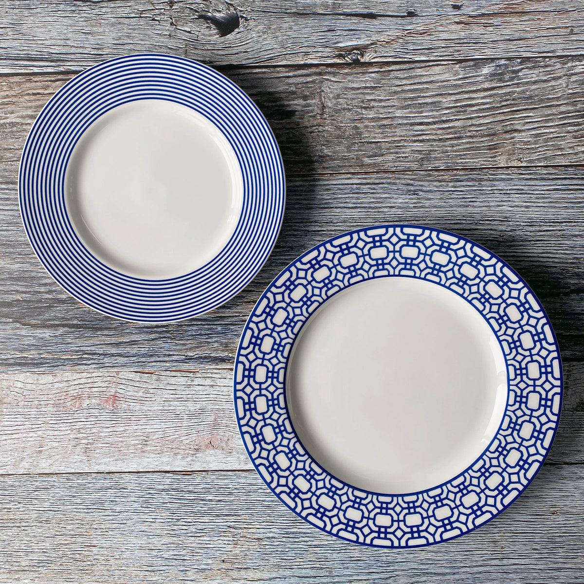 Two high-fired porcelain salad plates rest on a wooden surface. The left plate features the charming Newport Stripe Rimmed Salad Plate from Caskata Artisanal Home, while the right plate boasts a striking blue geometric pattern.