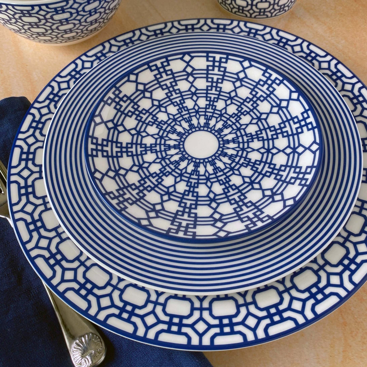 A set of blue and white plates with an intricate interlocking lattice pattern, stacked on a table beside a dark blue napkin and silverware, showcases the Newport Small Plates collection from Caskata Artisanal Home made from premium porcelain.