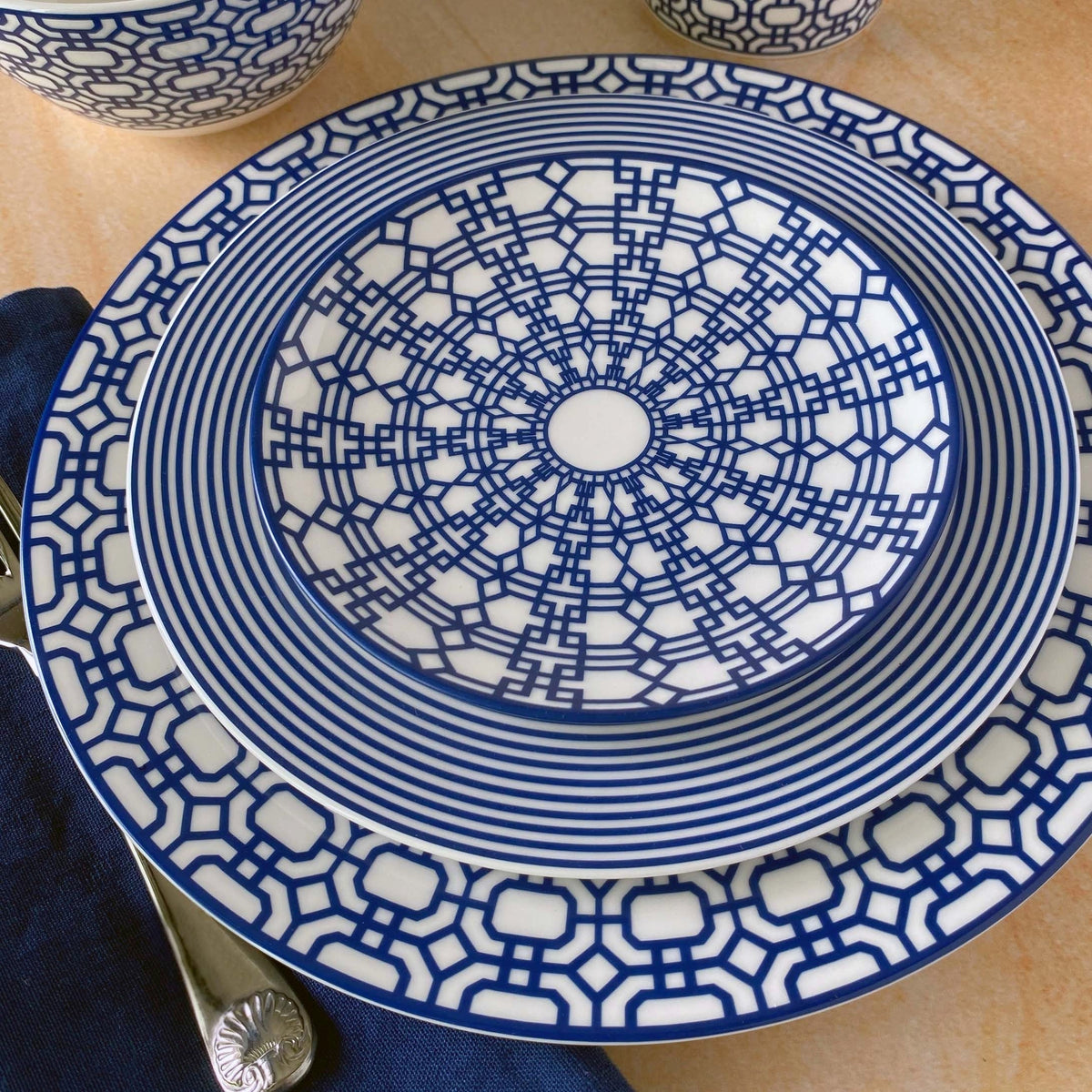 A Newport Garden Gate Rimmed Salad Plate from Caskata Artisanal Home with intricate blue geometric patterns on a light surface, reminiscent of the Newport Garden Gate design. A dark blue napkin and a silver spoon are partially visible in the bottom left corner, enhancing this premium porcelain display.