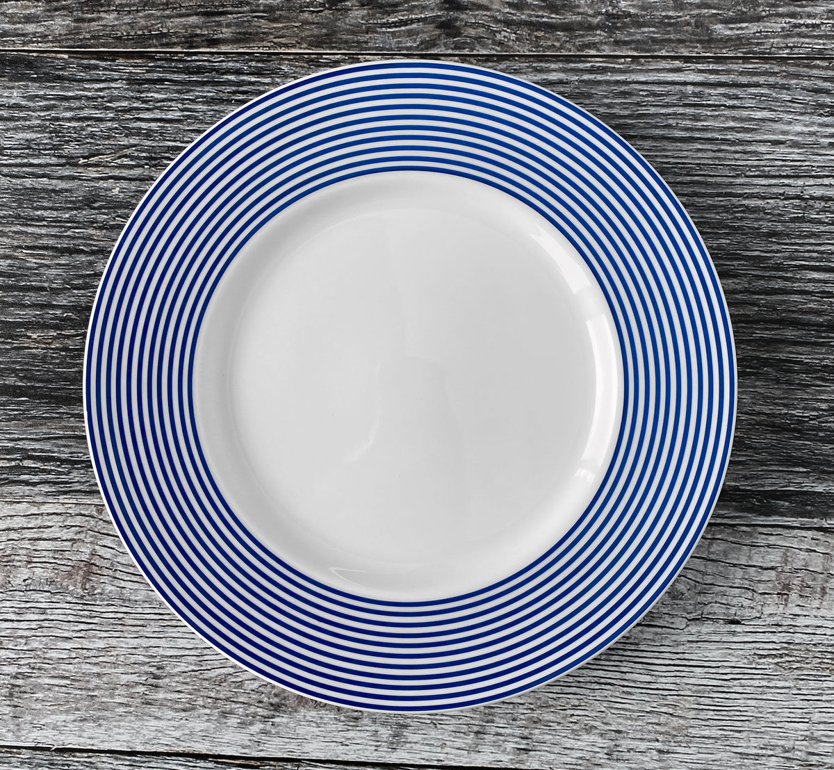 A Newport Stripe Rimmed Salad Plate from Caskata Artisanal Home, with multiple blue concentric circles around its edge, resembling the classic Newport Stripe, is placed on a weathered wooden surface.