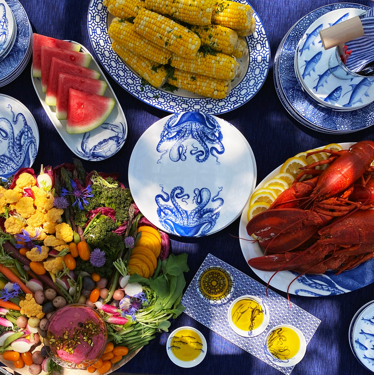 A contemporary blue and white Newport Garden Gate Oval Rimmed Platter table setting featuring a lobster, corn, and watermelon by Caskata Artisanal Home.