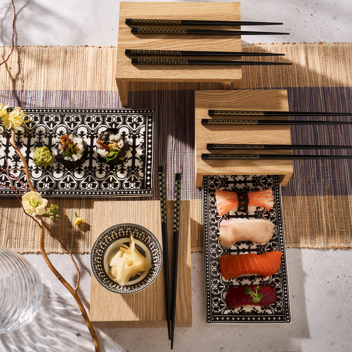 A neatly arranged Japanese sushi meal setup, featuring sushi rolls, sashimi, a bowl of pickled ginger on Caskata Casablanca Medium Sushi Trays, Set of 2, and chopsticks on patterned plates and wooden blocks, atop a straw mat background.
