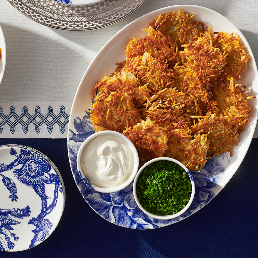 A plate of crispy latkes served with sour cream and chopped chives, on a Peony Medium Coupe Oval Platter from Caskata Artisanal Home, with an elegant blue and white dinnerware pattern.