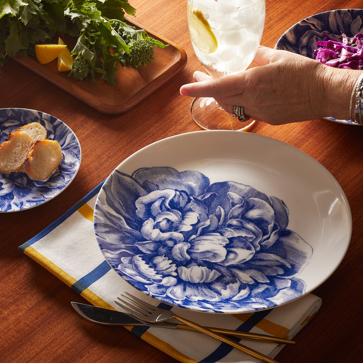 A person&#39;s hand placing a slice of pork tenderloin onto a premium porcelain Caskata Artisanal Home Peony Coupe Dinner Plate on a wooden table, with a drink and salad nearby.