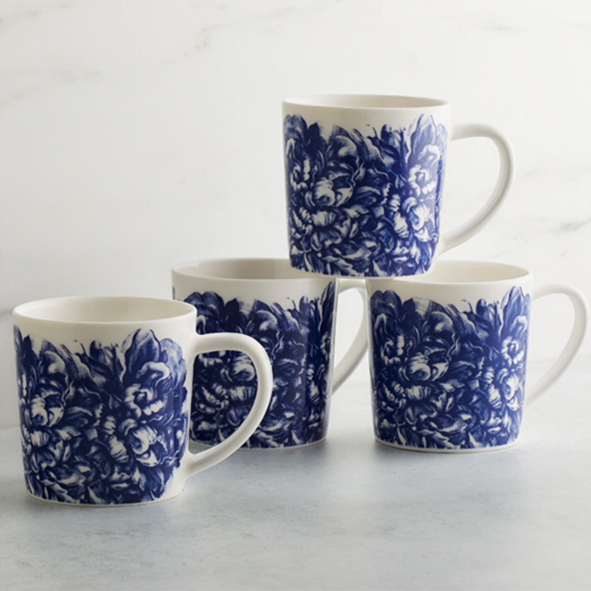 Four white Caskata Artisanal Home Peony Mugs with blue floral patterns stacked in two columns against a marble background.