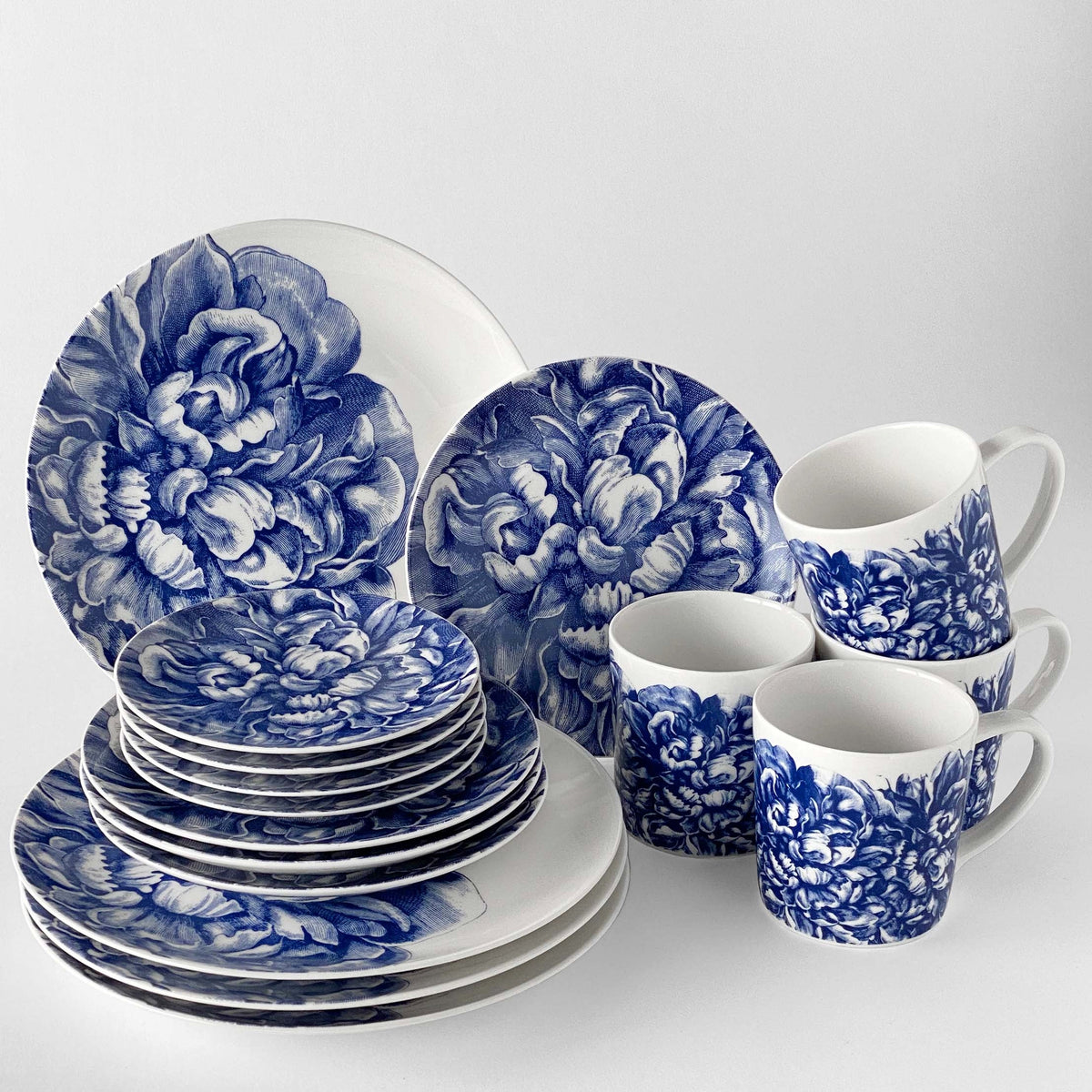 A set of Caskata Artisanal Home premium porcelain dinnerware, including Peony Coupe Dinner Plates and mugs, features a blue and white floral pattern displayed against a light background.