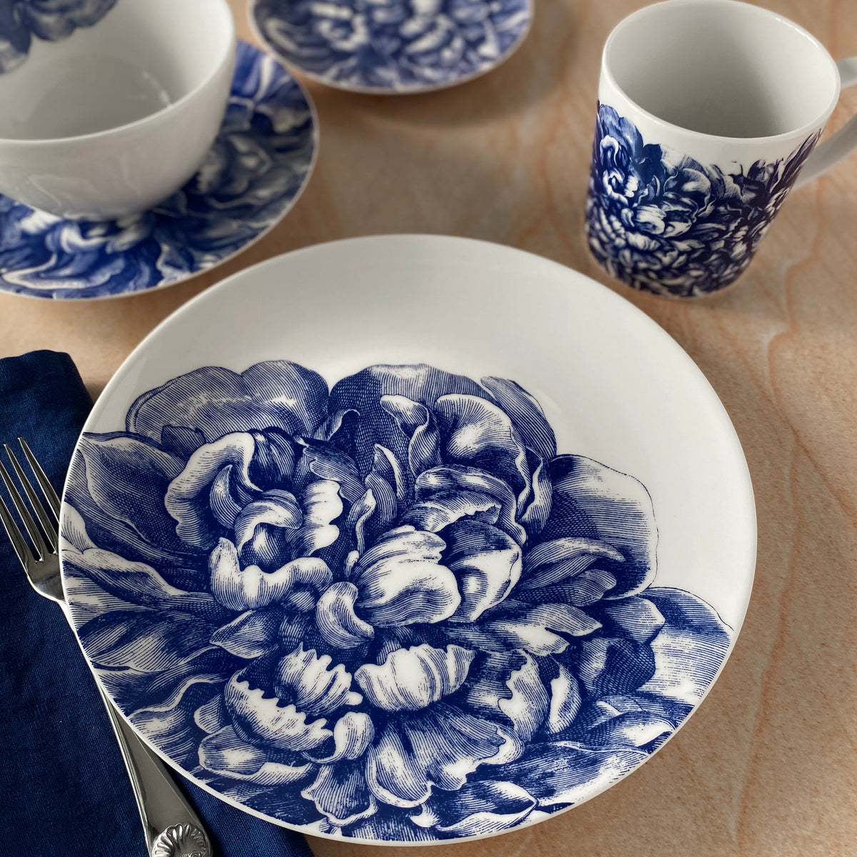 Elegant blue and white floral Caskata Artisanal Home Peony 4-Piece Place Setting including plates, bowls, and a cup, arranged on a table with a blue napkin and silverware.