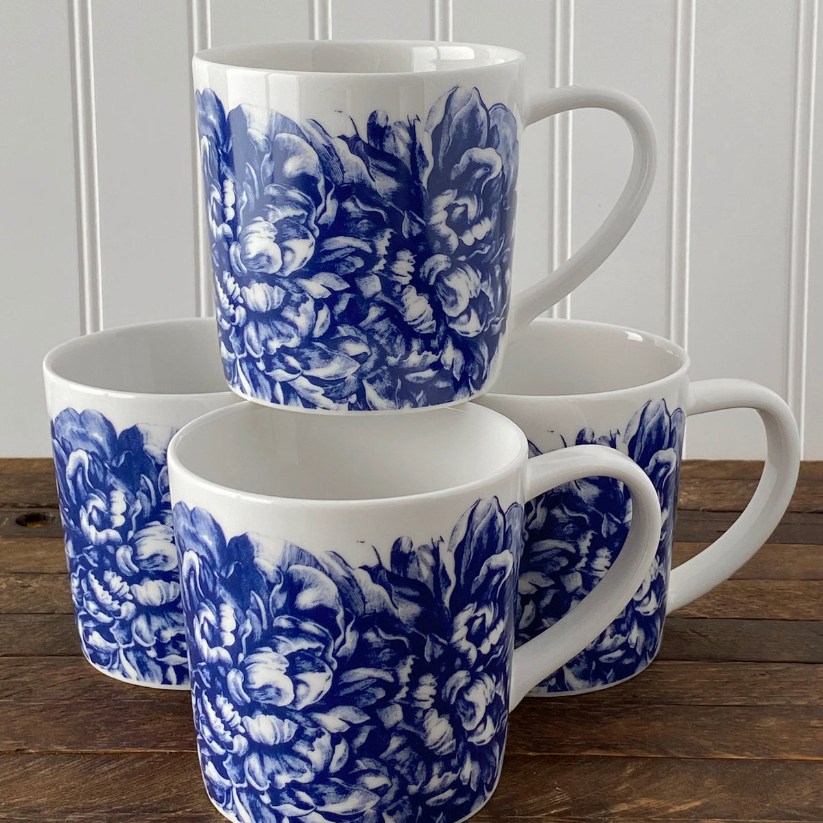 Four Peony mugs from Caskata Artisanal Home with blue floral patterns stacked in pairs on a wooden table, against a white beadboard background.