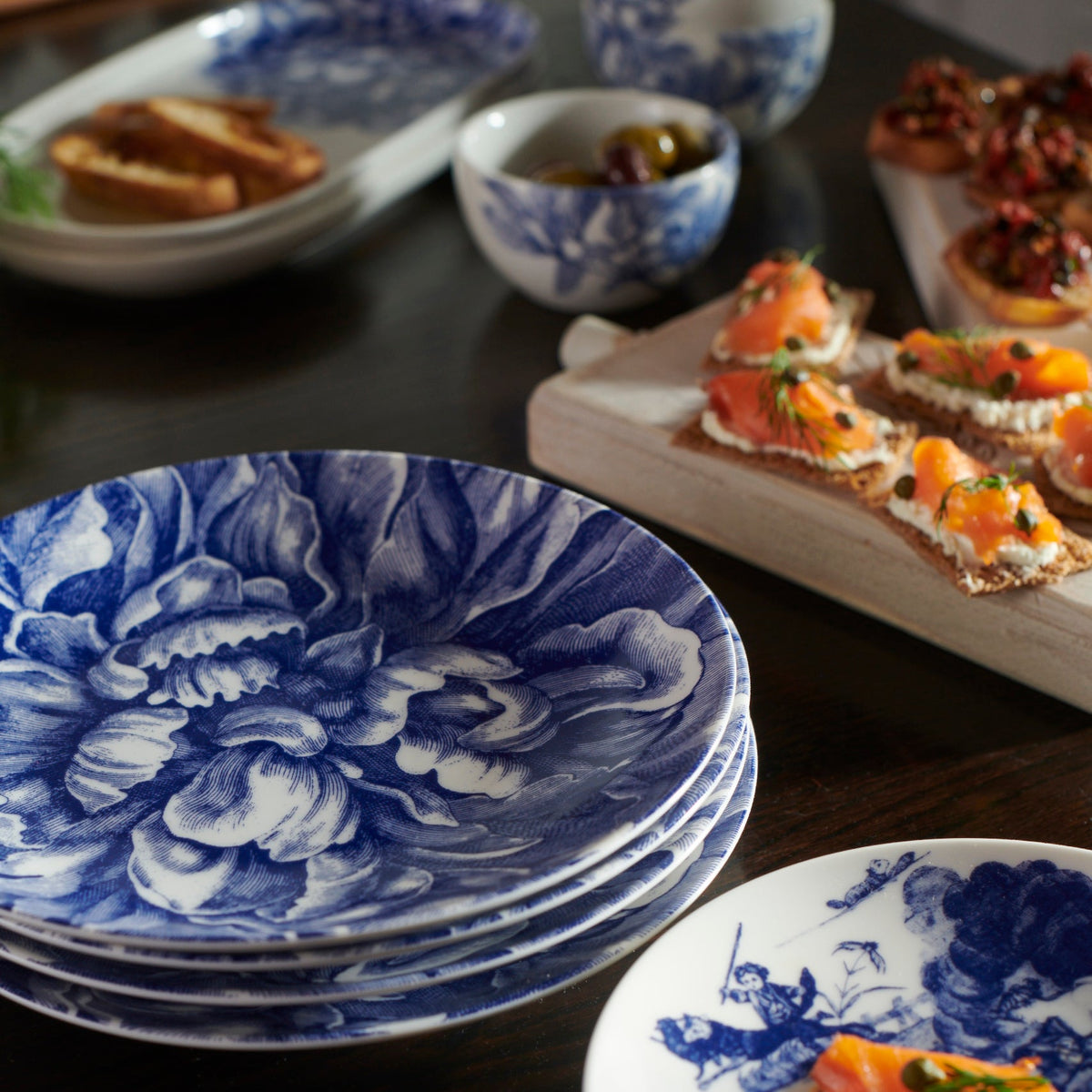 A table set with blue and white Peony 4-Piece Place Setting dinnerware from Caskata Artisanal Home, featuring premium porcelain plates and a variety of appetizers, including canapés and small bowls of sauce.