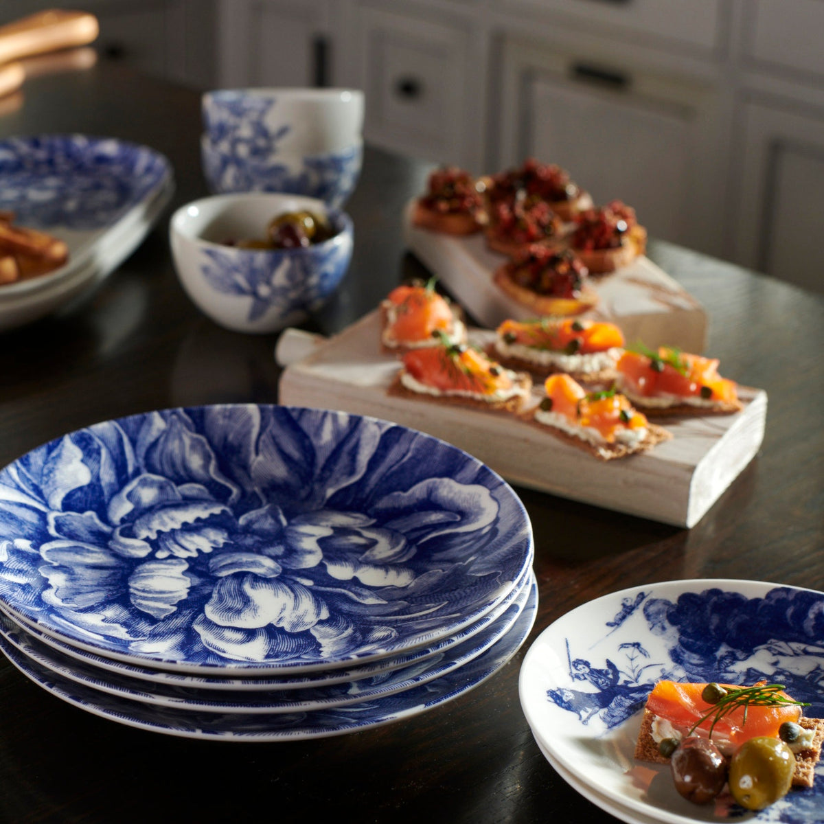 A dining table set with a selection of appetizers on wooden boards and two hands reaching for food, with premium Caskata Artisanal Home Peony Coupe Salad Plates in the foreground.