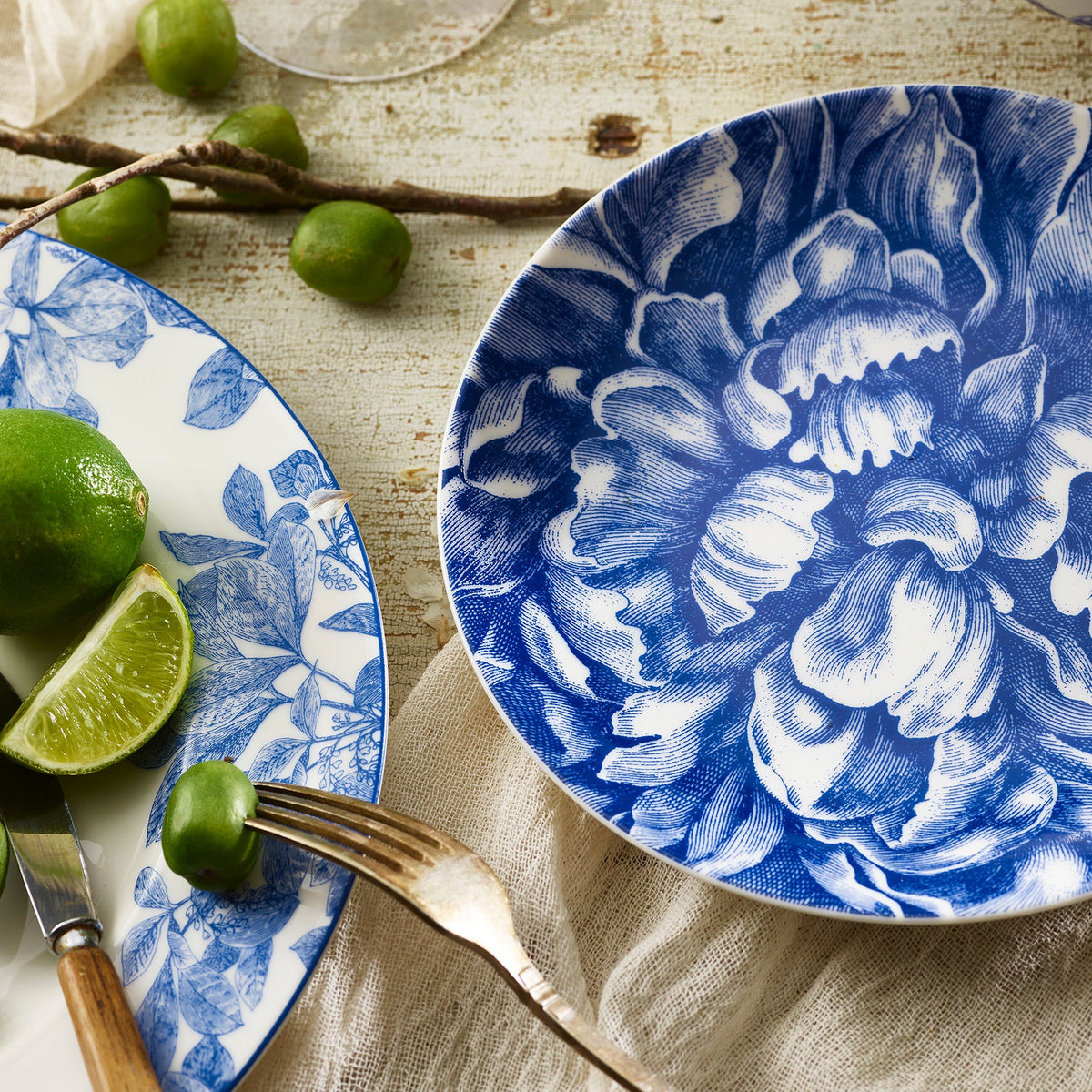 Blue and white floral Caskata Artisanal Home Peony Coupe Salad Plates with whole and sliced limes, and vintage silverware on a wooden table.