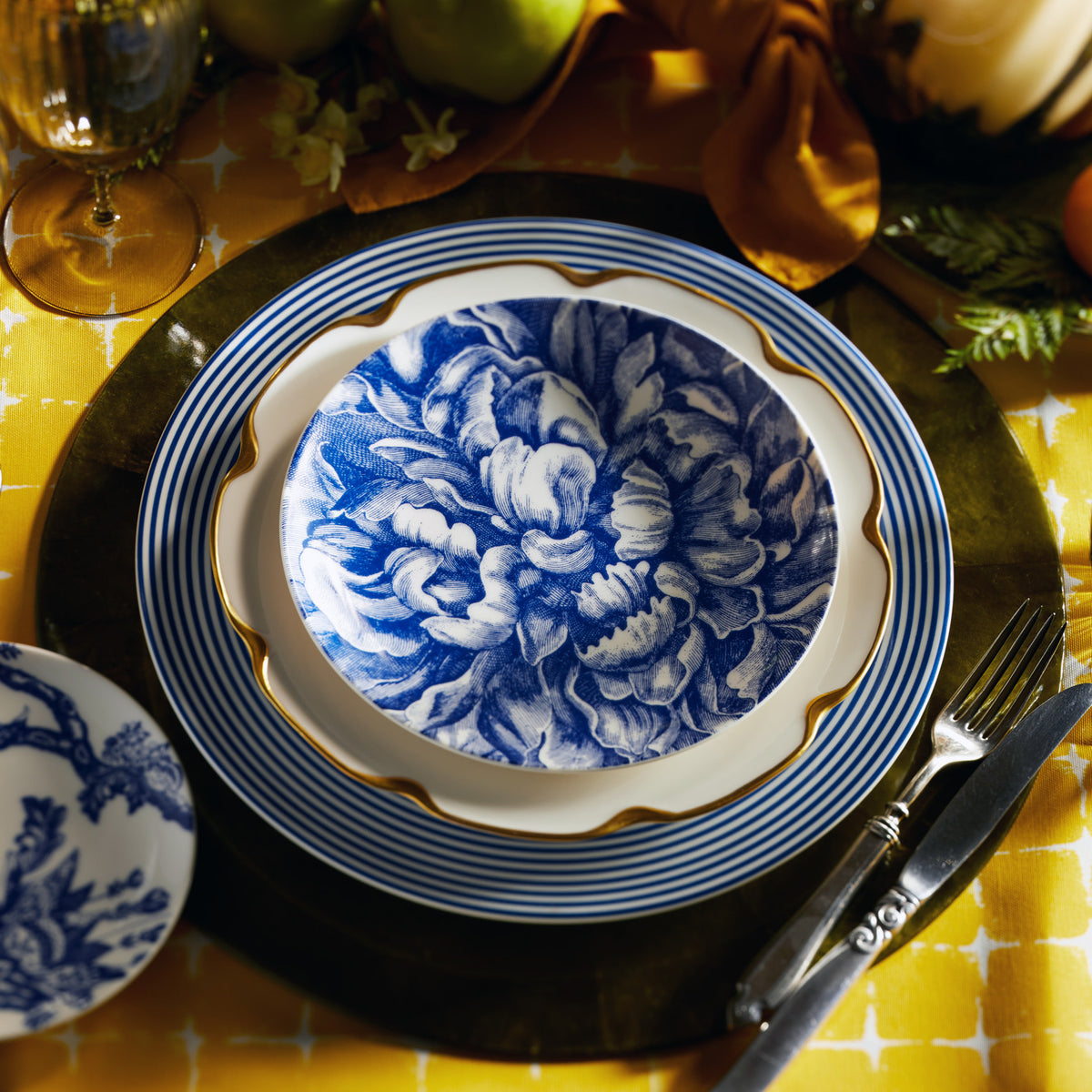 A table setting with a blue floral-patterned plate on top of a gold-rimmed white high-fired porcelain plate, all placed on a dark Newport Rimmed Charger Plate by Caskata Artisanal Home with a fork and knife on the right side. Yellow tablecloth in the background.