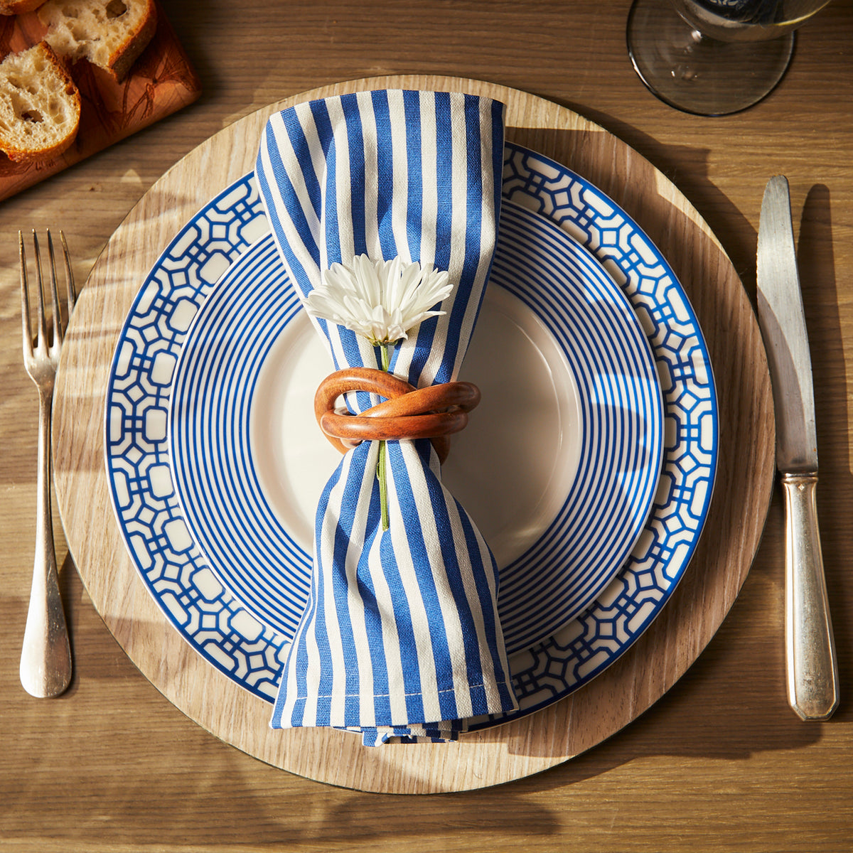 A place setting with a porcelain Newport Garden Gate Rimmed Dinner Plate by Caskata Artisanal Home featuring a blue and white pattern, striped napkin with a white flower, utensils, and a wooden placemat. Bread slices are in the top left corner, adding to the coastal collection ambiance.