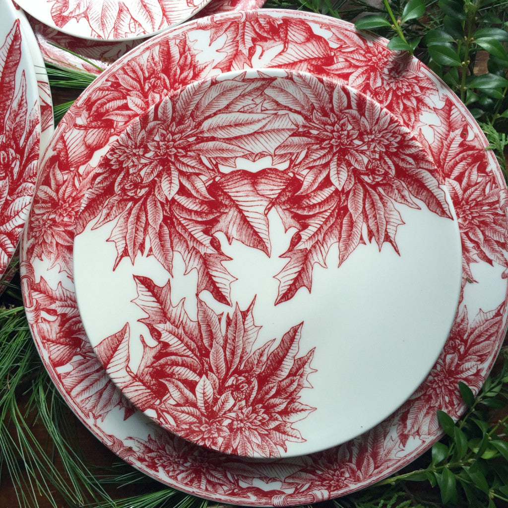 A set of Poinsettia Coupe Salad Plates by Caskata Artisanal Home, adorned with intricate red poinsettia floral patterns, graces a holiday table on a wooden surface with greenery in the background.