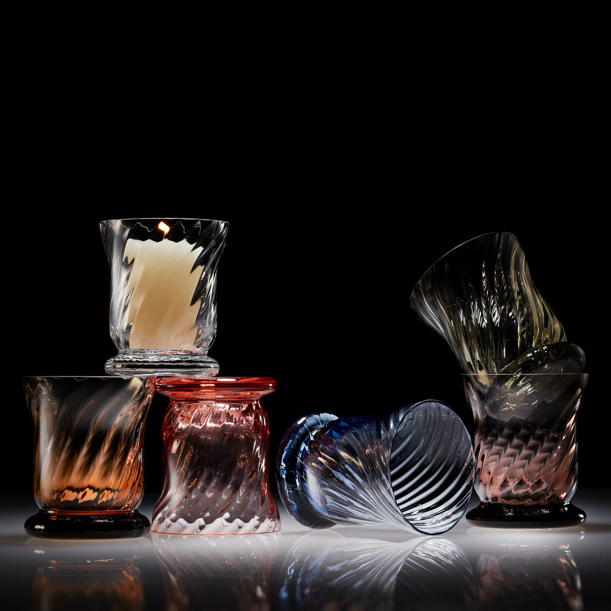 Caskata&#39;s collection of Quinn Votives are playfully stacked in front of a black background. Amber, Clear, Rose, Ocean, Smoke and Mocha Votives showcase the colorful line of glassware. The Clear Votive features a lit votive candle.