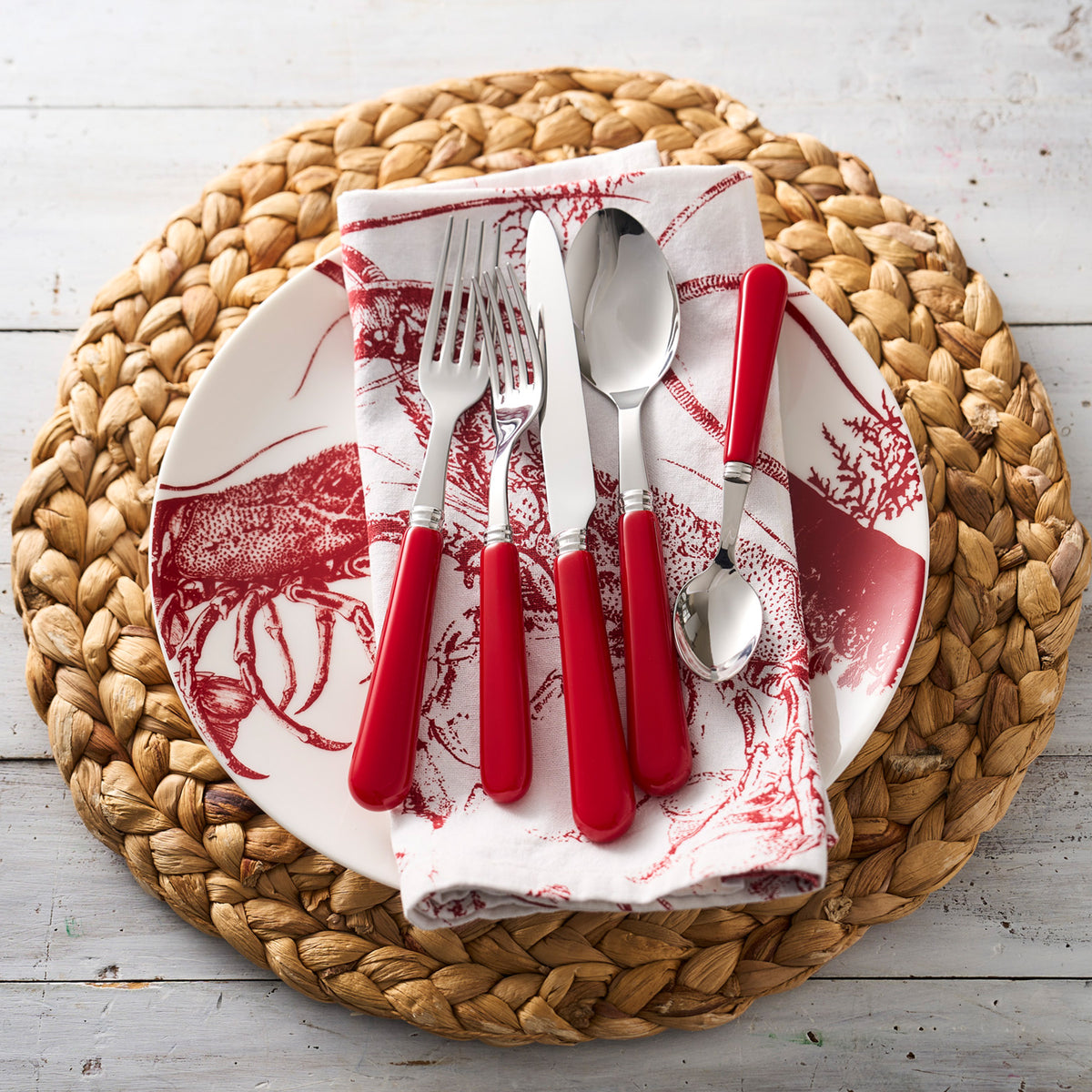 A place setting with red-handled cutlery and a red patterned napkin on a white seaside style Lobster Coupe Dinner Plate by Caskata, seated elegantly on a woven placemat.