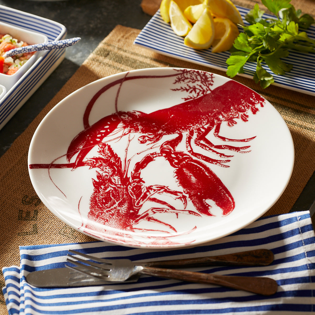 A Caskata Lobster Coupe Dinner Plate made of premium porcelain features a red lobster illustration on a white backdrop, placed on a table with a striped blue and white napkin, fork, and knife. The plate is dishwasher and microwave safe. Lemon wedges and greens add to the coastal charm in the background.