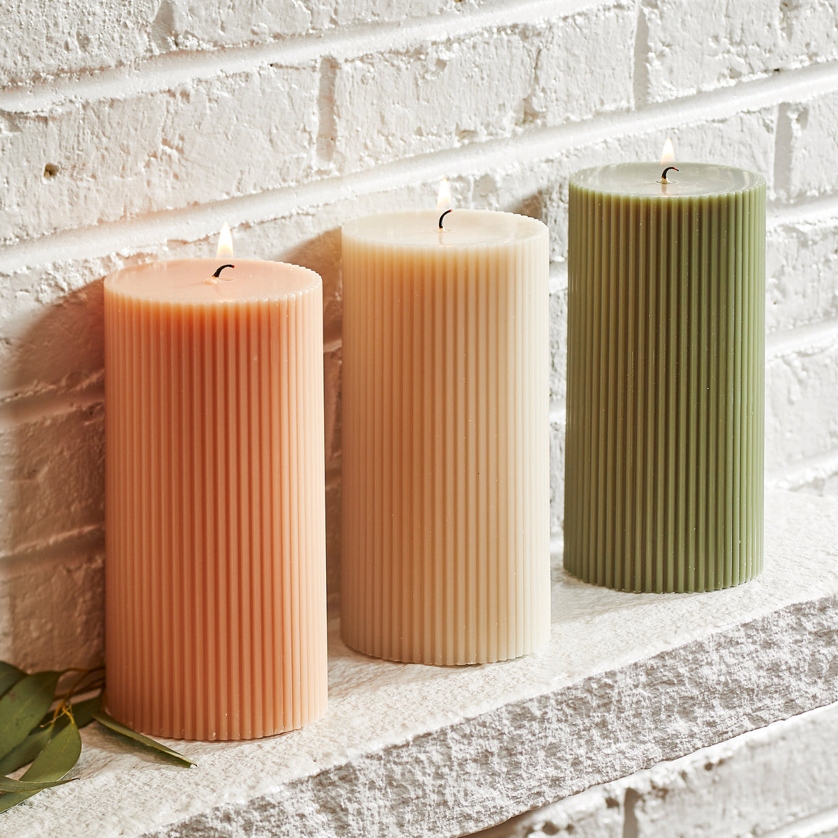 The ribbed Pillar Candle in Petal Pink 6 inch from Caskata is also available in ivory white and moss green. The set of three candles is displayed on a brick mantel.