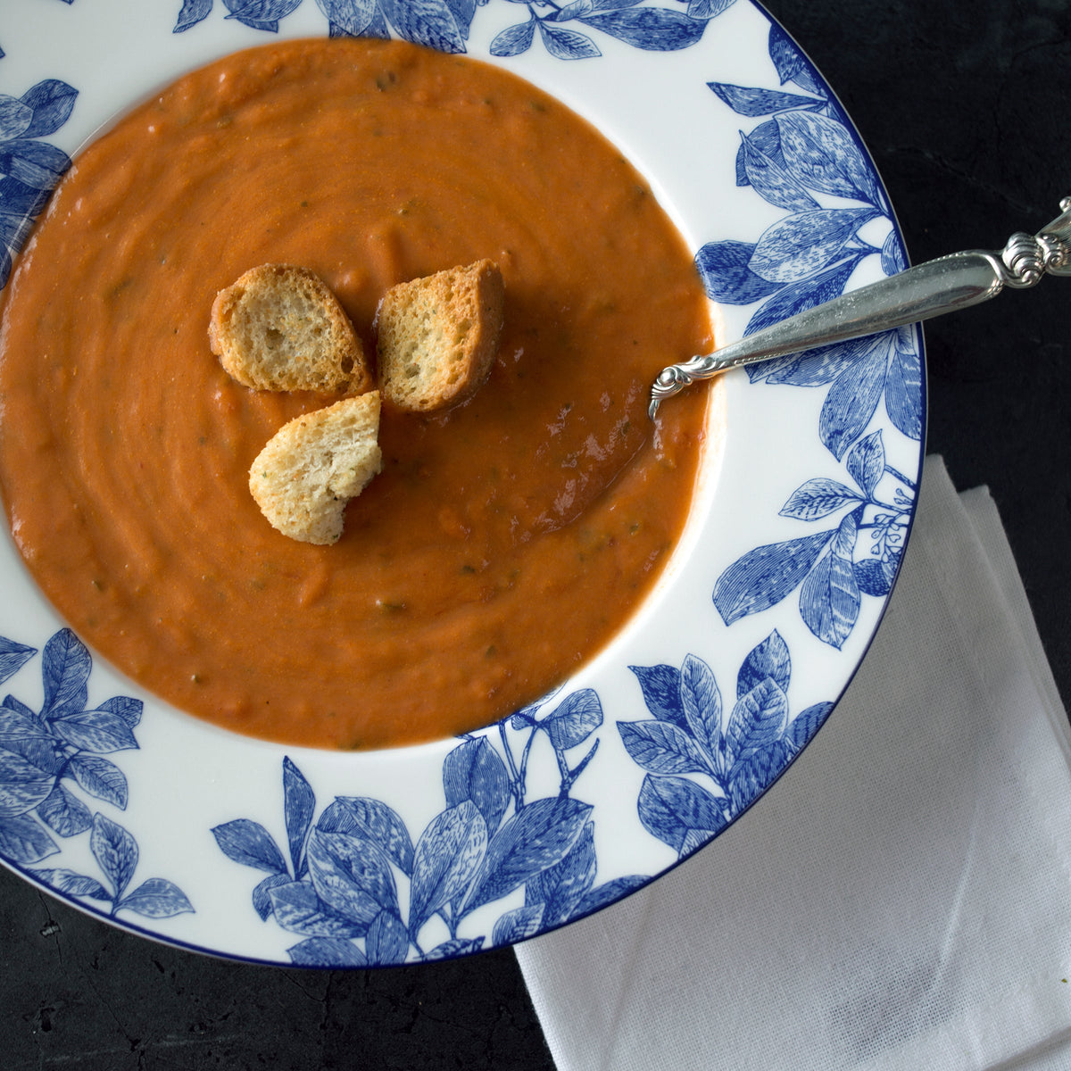An Arbor Rimmed Soup Bowl of high-fire porcelain from Caskata Artisanal Home cradles a savory tomato soup with three croutons, served on a white plate with blue floral patterns. A silver spoon rests on the edge next to a white napkin. The entire setting is dishwasher safe for easy cleanup.