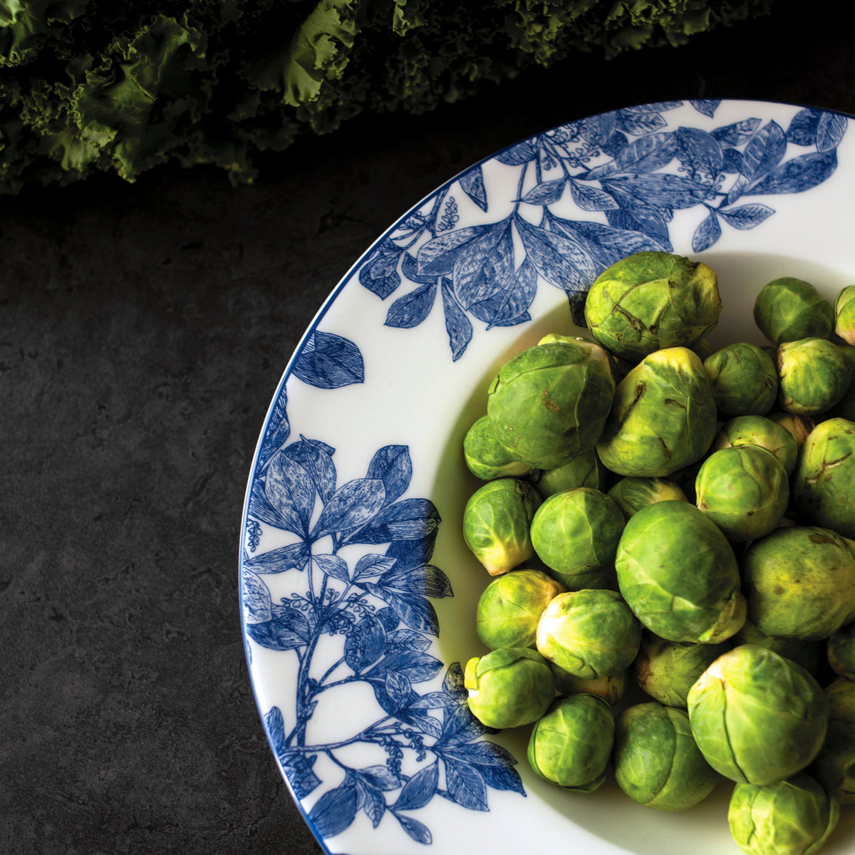 A white Arbor Rimmed Soup Bowl with blue floral patterns, crafted from high-fire porcelain, holds fresh green Brussels sprouts on a dark surface.