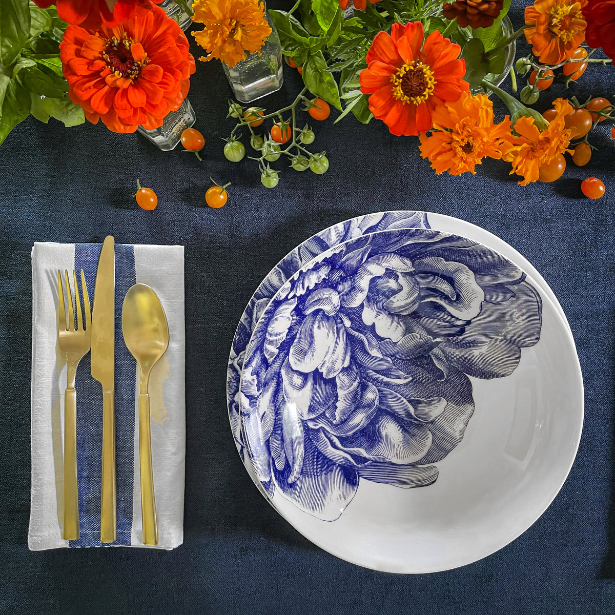 A table setting featuring Peony Coupe Soup Bowls from Caskata Artisanal Home in blue floral, gold cutlery, and bright orange flowers on a dark blue tablecloth.