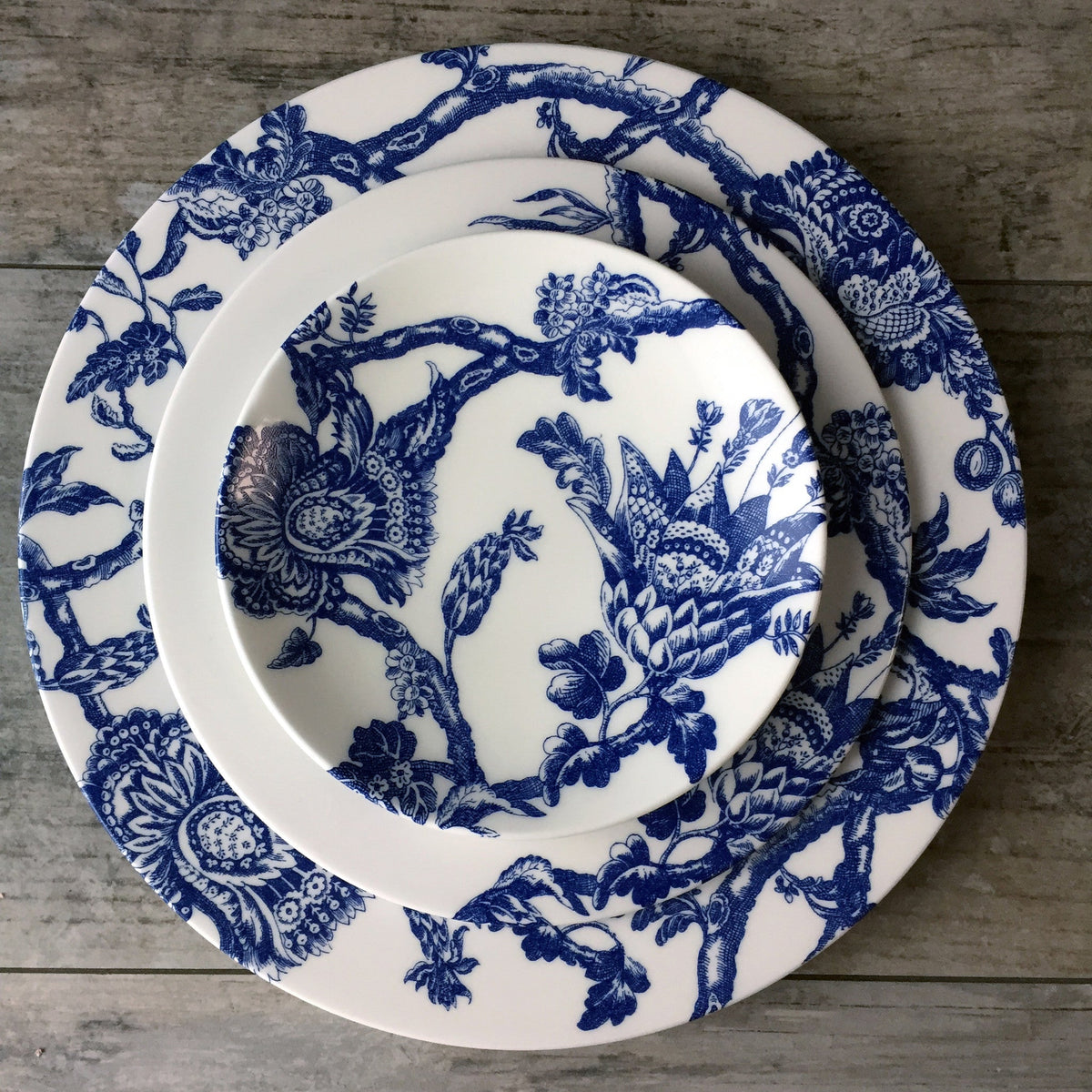 Three stacked white Arcadia Rimmed Salad Plates with intricate blue floral patterns on a gray wooden surface, crafted from Caskata Artisanal Home dinnerware inspired by the Williamsburg Foundation collection.