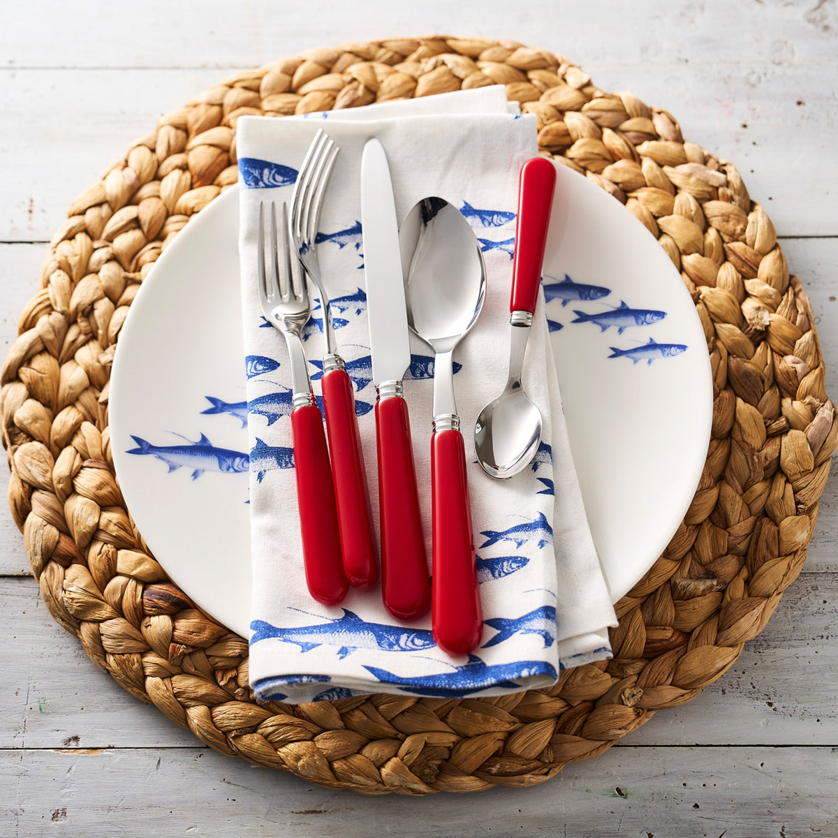A neatly arranged dinner setting with School of Fish Coupe Dinner Plates from Caskata Artisanal Home, red-handled cutlery on a blue and white napkin, all resting on a round woven placemat.