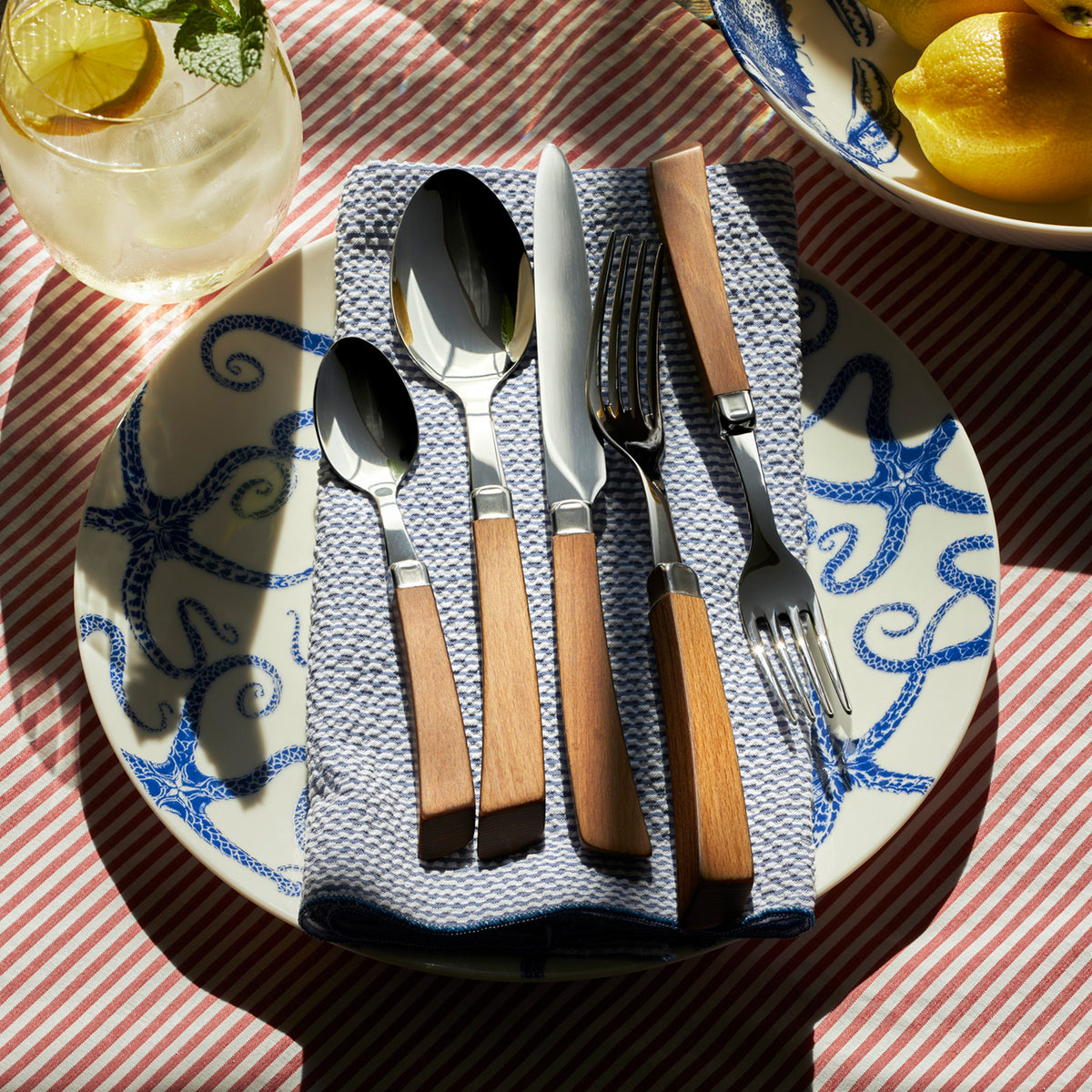 A table setting with wooden-handled cutlery on a Starfish Coupe Dinner Plate by Caskata Artisanal Home, laid on a blue towel. A drink with lemon and a bowl of lemons are also visible on a red striped tablecloth, creating a charming coastal vibe reminiscent of starfish-dotted shores.