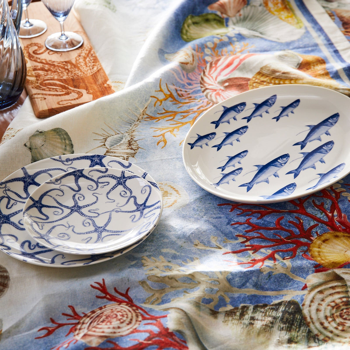 A tabletop featuring a cloth with sea life designs, and a set of white premium porcelain plates with blue sea creatures—one with fish and one with octopuses. Three wine glasses and a wooden board are partially visible, alongside a charming Starfish Coupe Salad Plate by Caskata Artisanal Home.