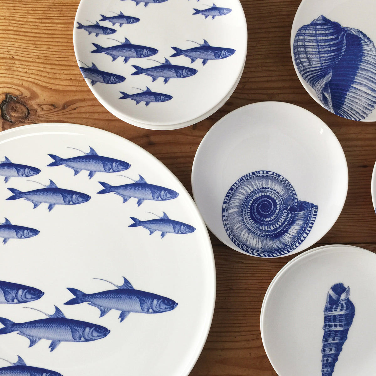 A collection of heirloom-quality dinnerware featuring blue fish and shell designs is displayed on a wooden surface. The Caskata Artisanal Home School of Fish Small Plates are expertly arranged, showcasing their intricate and captivating patterns.