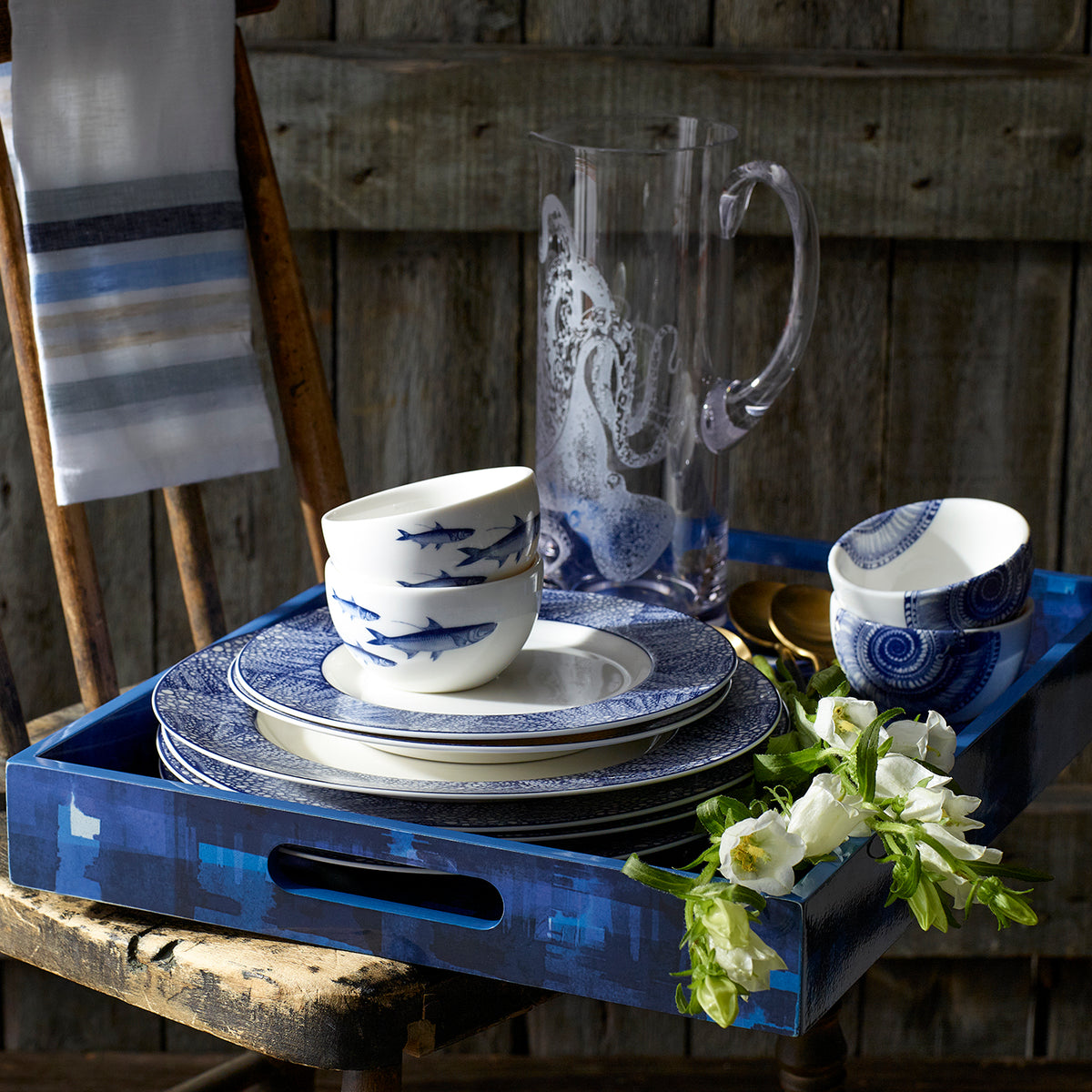 A Caskata Artisanal Home wooden tray with a Shells Snack Bowl and a pitcher among blue and white dishes, reminiscent of a shell treasure.