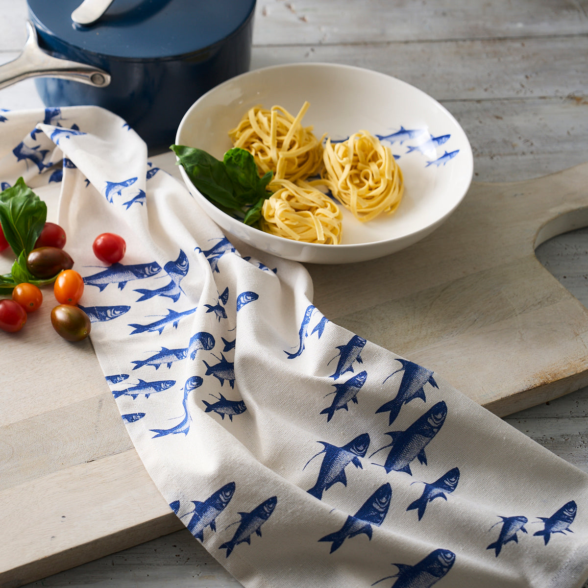 A blue pot and a white plate containing uncooked pasta and basil rest on a wooden surface, accompanied by cherry tomatoes, basil, and a charming school of fish-patterned cloth. This elegant setup features our School Fish Entrée Bowl from Caskata Artisanal Home, perfect for your culinary creations.