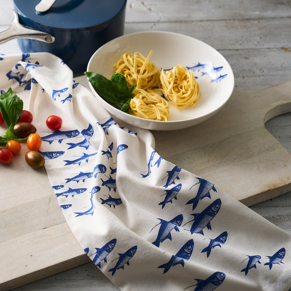 A bowl of pasta on a wooden table with a Caskata School of Fish Kitchen Towel, Set of 2, cherry tomatoes, basil, and a cooking pot in the background.