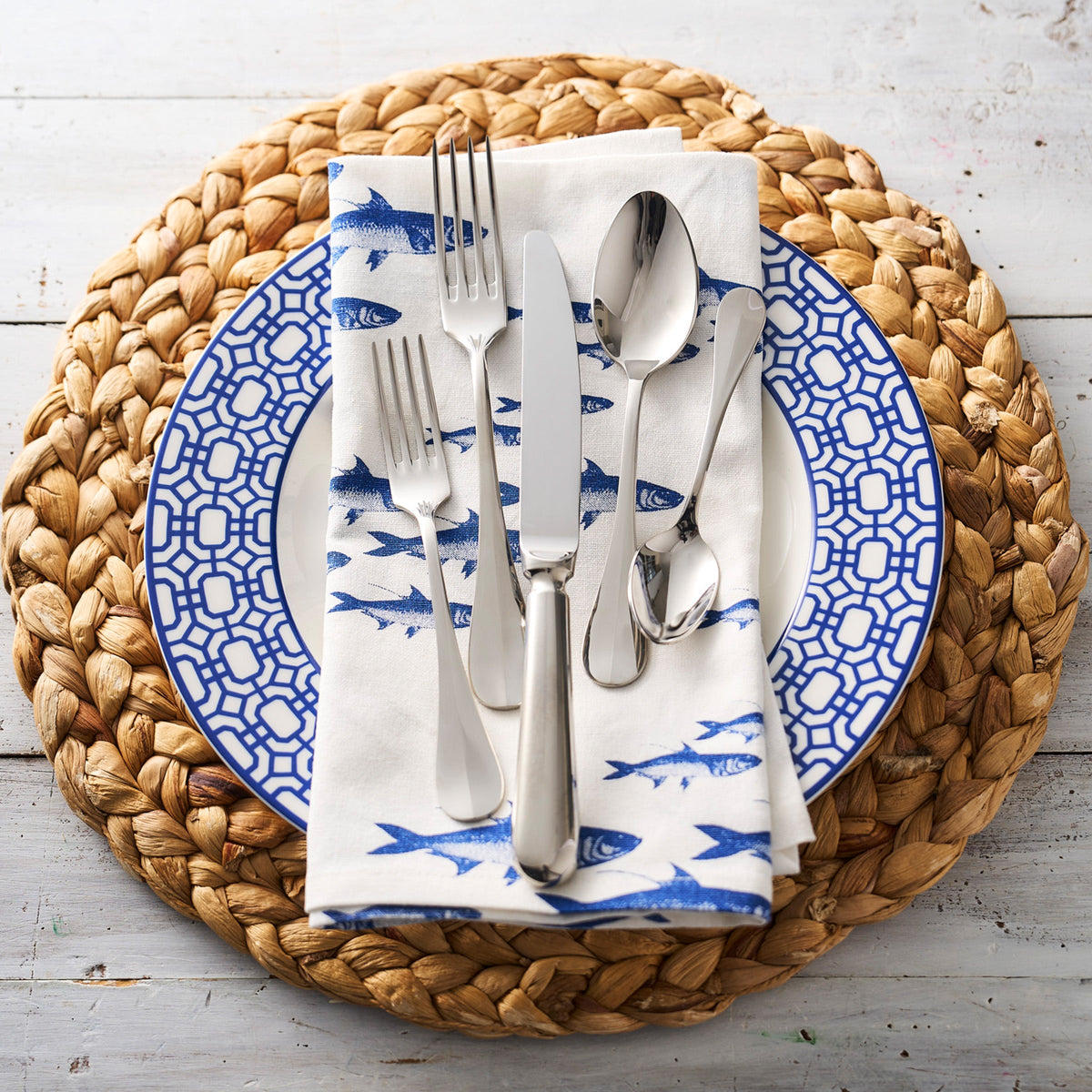Blue and white patterned plate from the Caskata School of Fish dinnerware collection, with School of Fish Dinner Napkins, set with silverware on a round woven placemat.