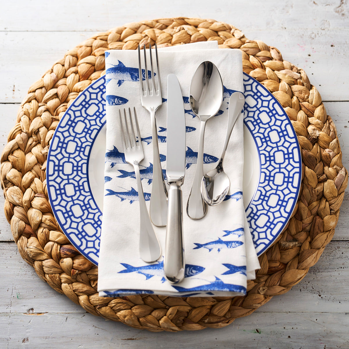 A woven placemat with a Caskata Artisanal Home Newport Garden Gate Rimmed Dinner Plate, a fish-patterned napkin, and silverware, including a knife, fork, and spoons, arranged neatly on a wooden surface - perfect for any coastal collections.