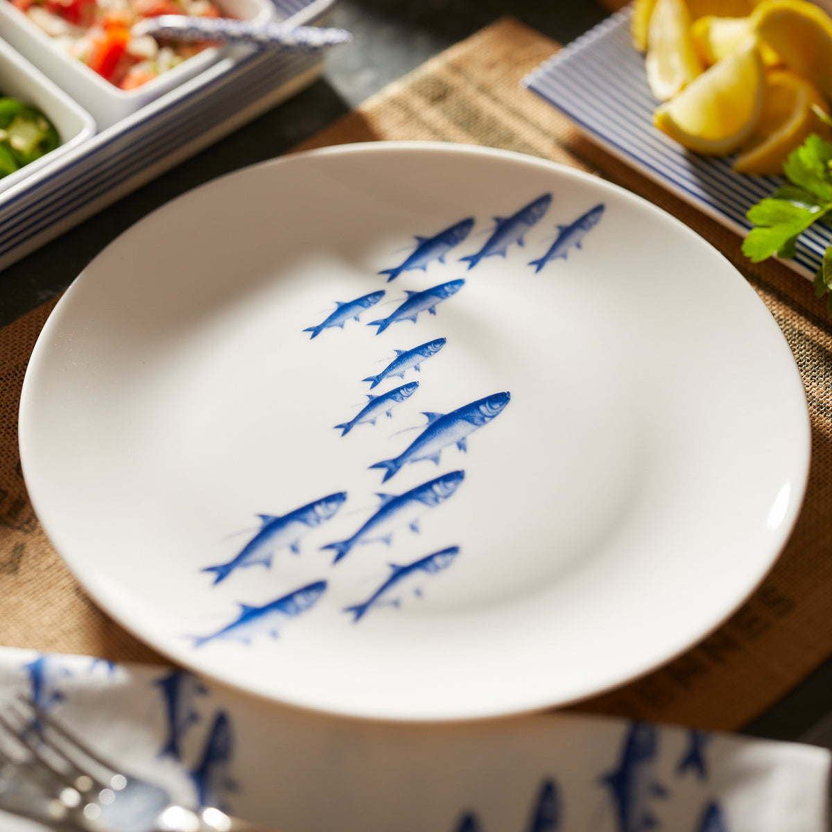 A School of Fish Coupe Dinner Plate from the Caskata Artisanal Home premium porcelain collection, surrounded by condiments and lemon wedges on a wooden table.