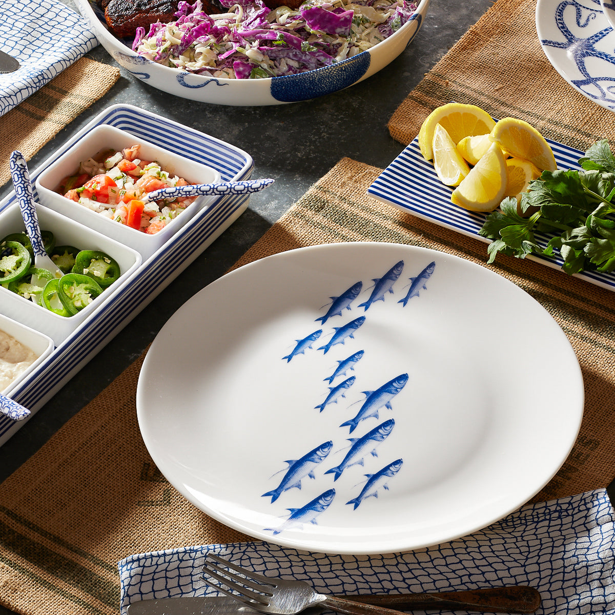 A School of Fish Coupe Dinner Plate with a blue fish pattern, surrounded by colorful dinnerware and sliced lemons, arranged on a textured tabletop from Caskata Artisanal Home.