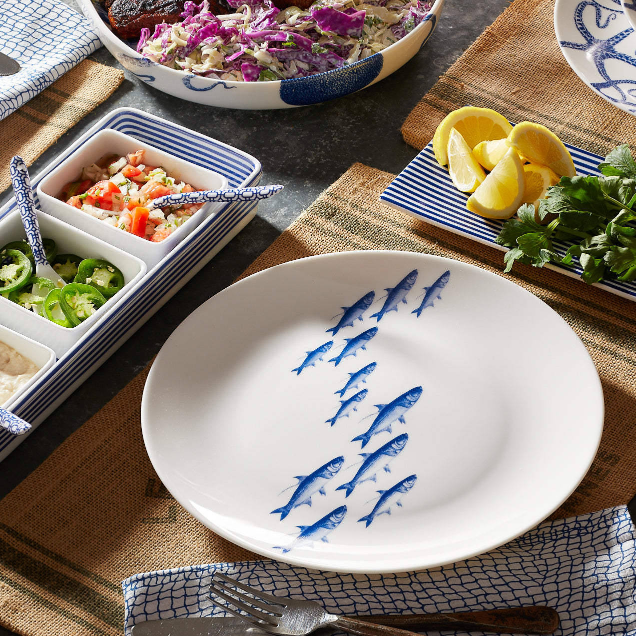 School of Fish Coupe Dinner Plate from Caskata Artisanal Home, a white ceramic plate with a pattern of blue fish swimming in a curved line, viewed from above; this premium porcelain dinnerware is dishwasher safe.